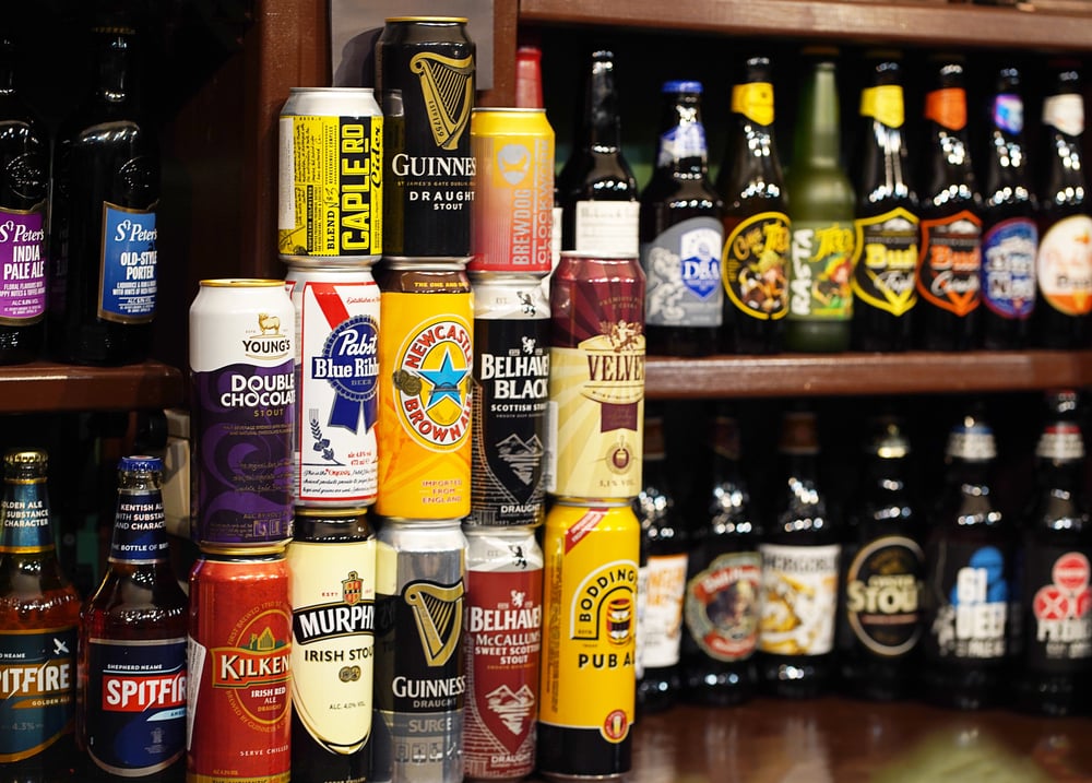 Stacked mix of craft beer cans and bottles in front of a packed bar shelf.