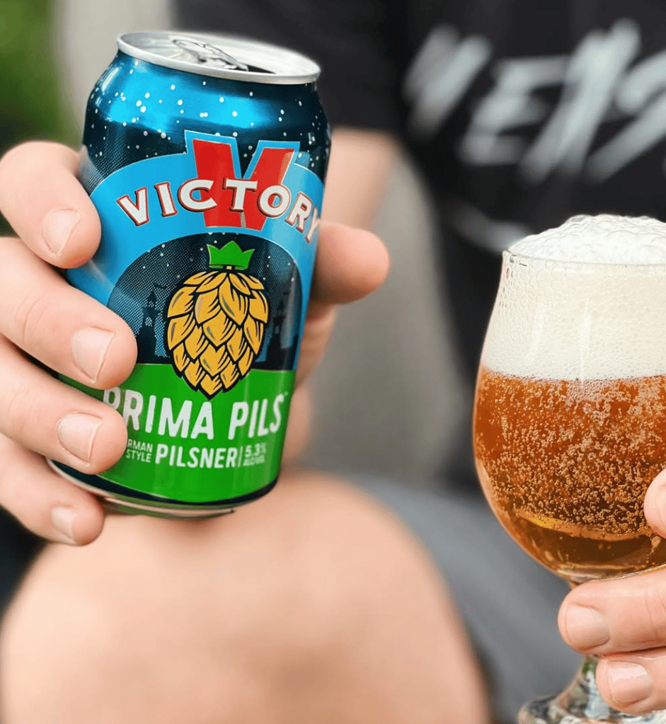 A man holding a Victory Prima Pils can in one hand and a beer glass in the other.
