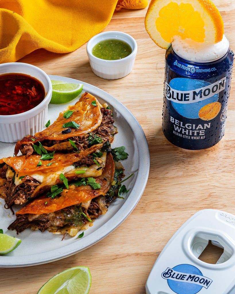 blue moon belgian white can with orange slice on rim next to birria tacos and branded bottle opener