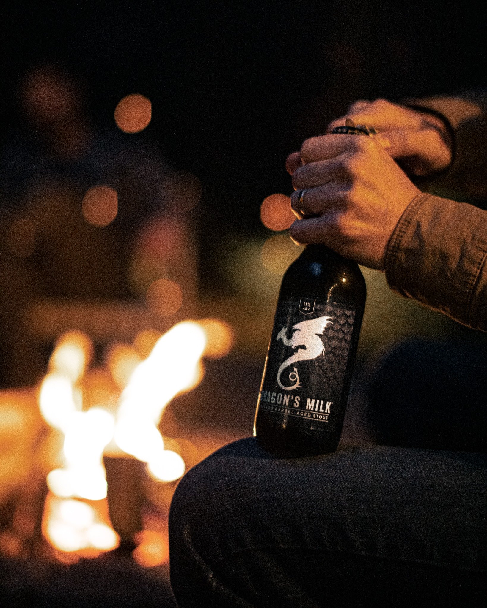 New Holland Dragon’s Milk bottle being opened outdoors next to a bonfire.