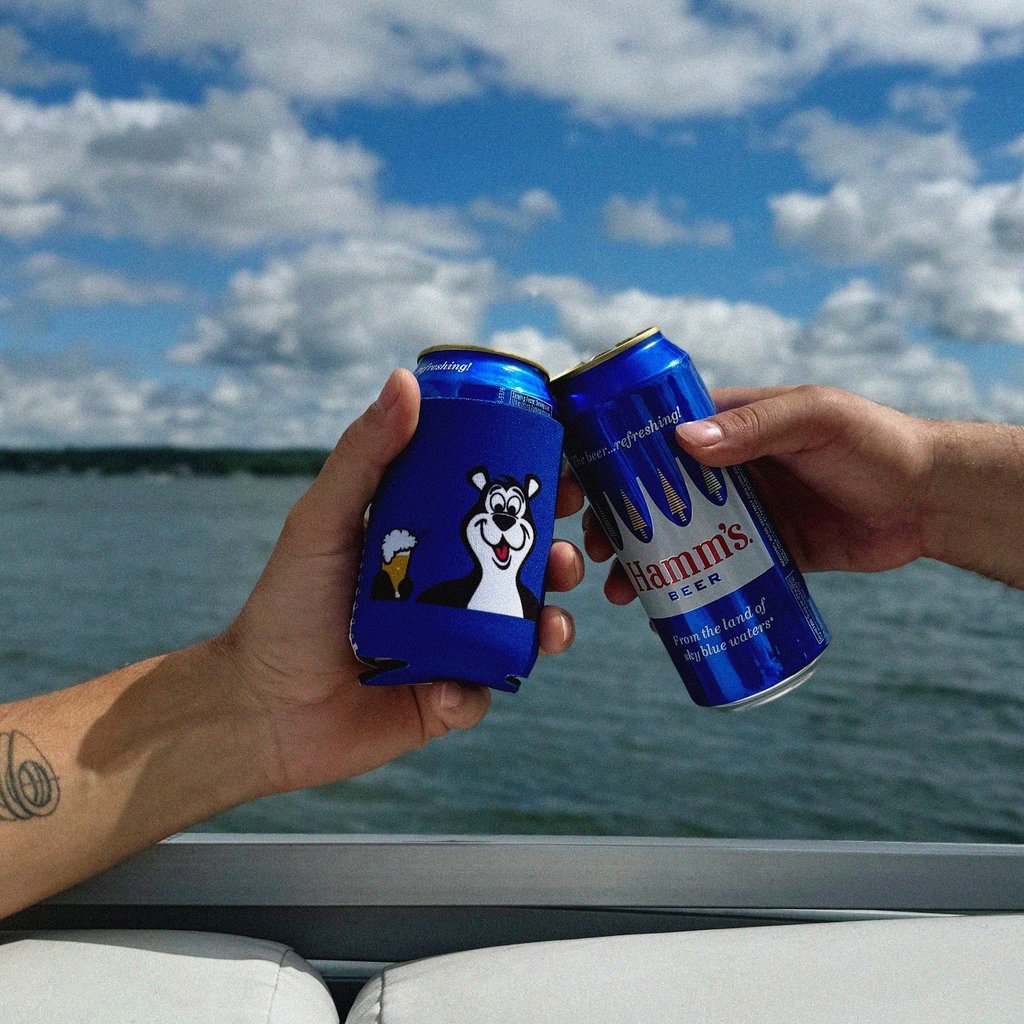 hamm's cans one in koozie being cheersed near a body of water