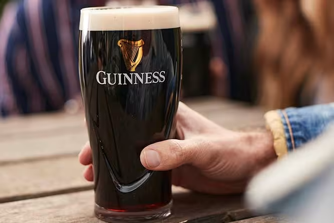 Hand holding a pint of dark Guinness Draught, the best cheap dark beer, with creamy head on a wooden table.