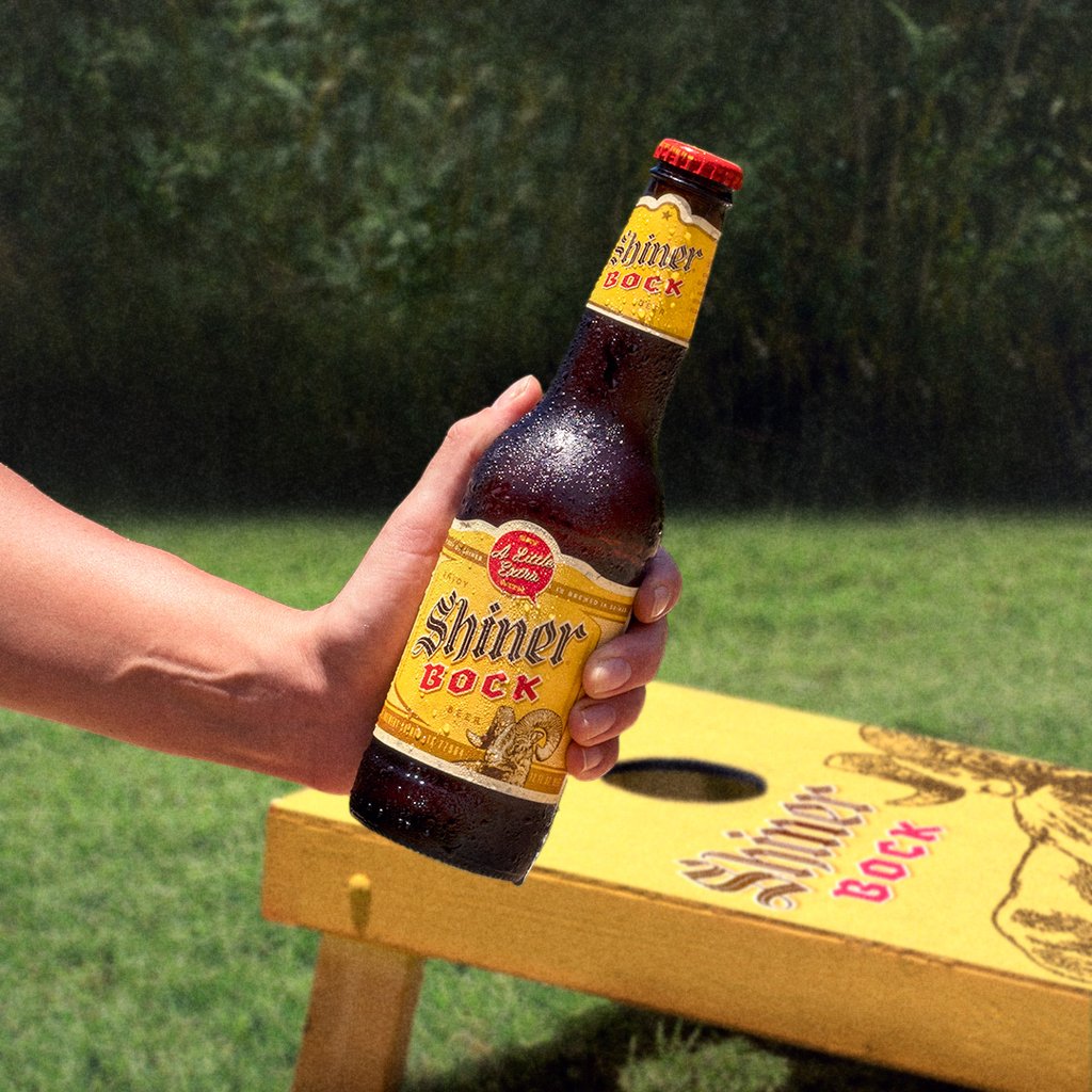 Shiner Bock bottle in hand beside a branded cornhole board