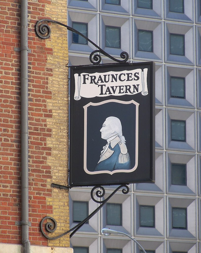 The sign for Fraunces Tavern, featuring an illustration of George Washington, with a skyscraper in the foreground.