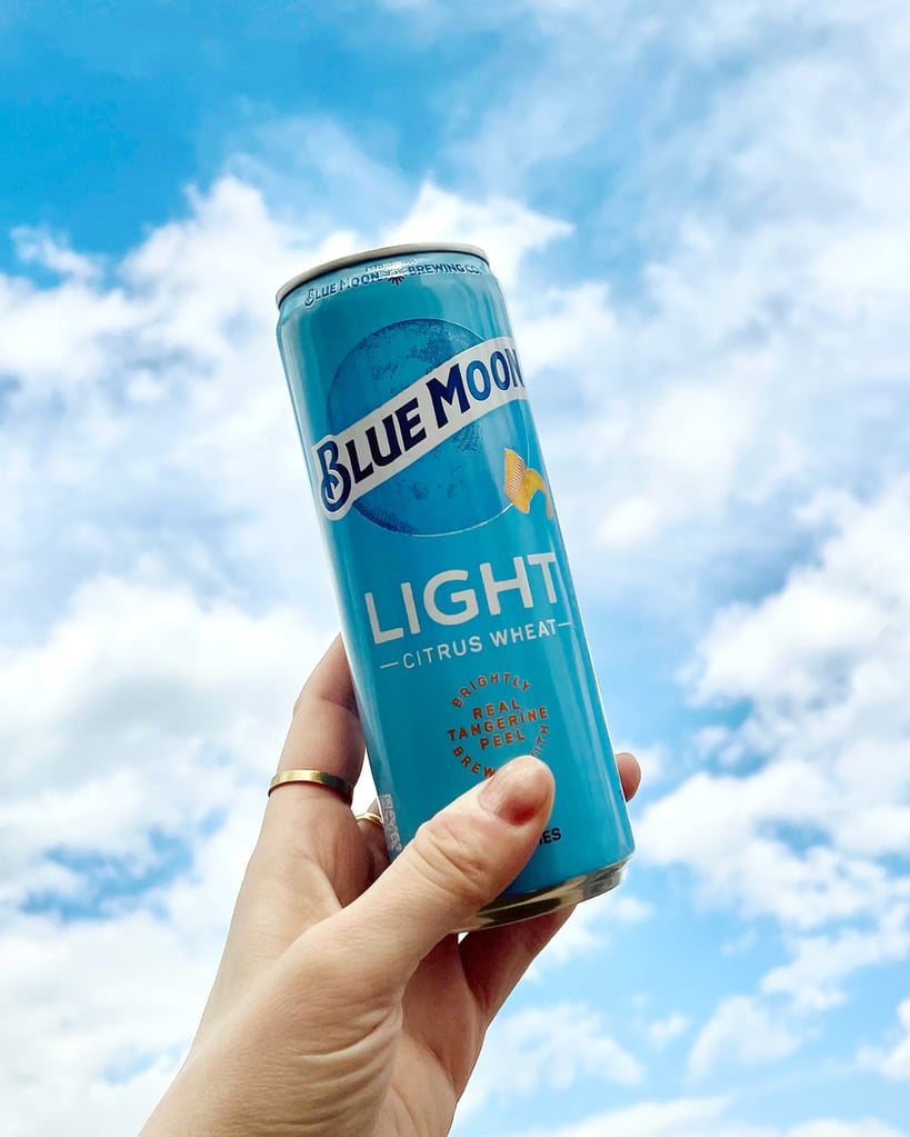 Blue Moon Light Citrus wheat can being held aloft in front of a cloudy sky