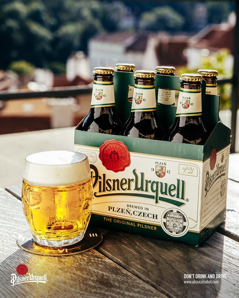 Pilsner Urquell six-pack of bottles with branded glassware on table.