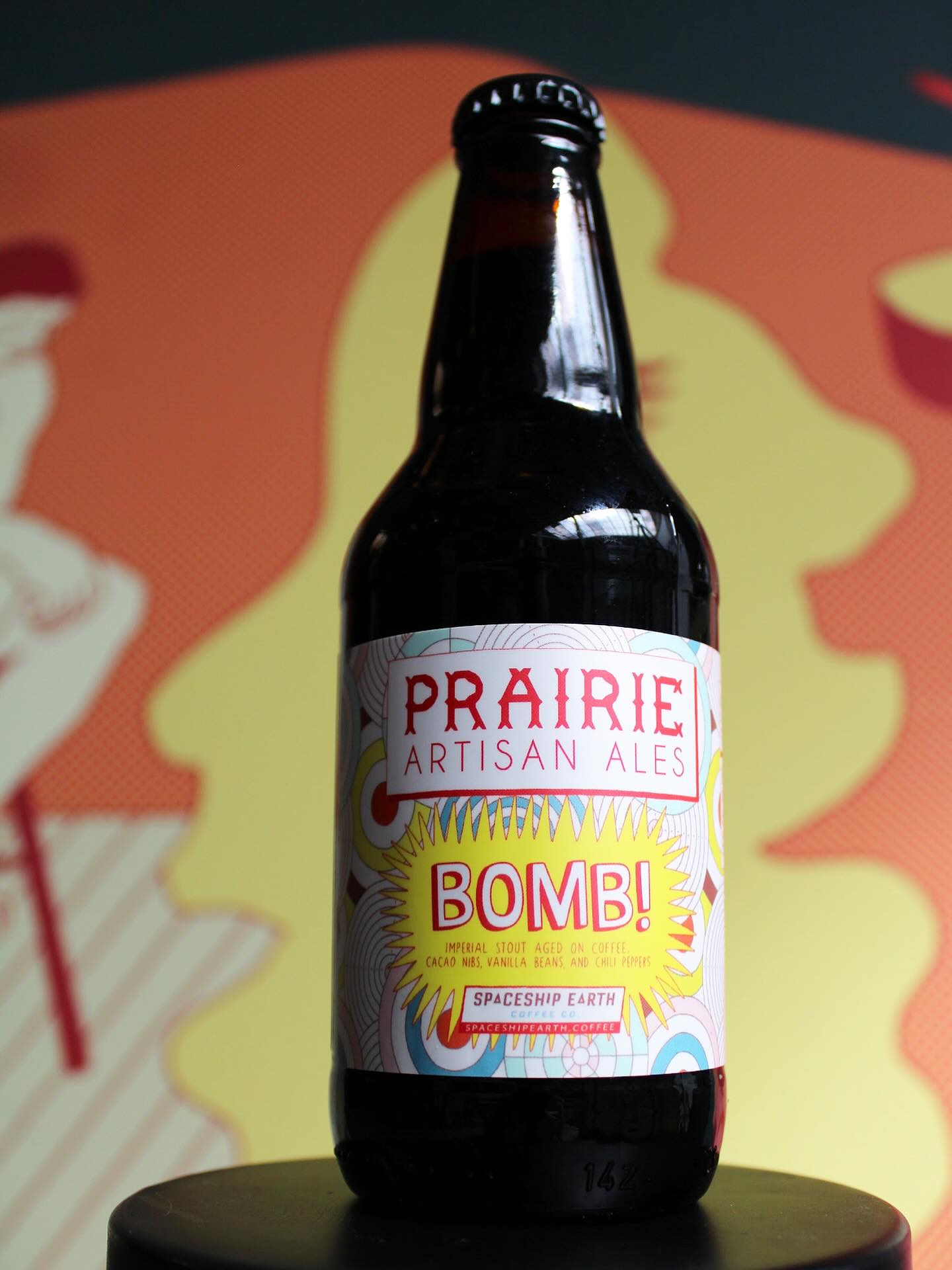Prairie Artisan Ales Bomb! bottle resting on stool in front of colorful wall.