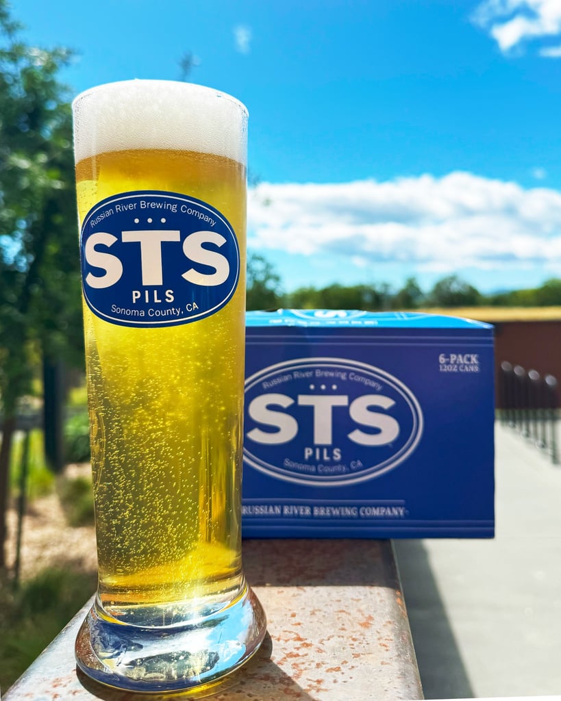 Russian River Brewing Co. STS Pils six-pack and branded glassware outside on a sunny day.