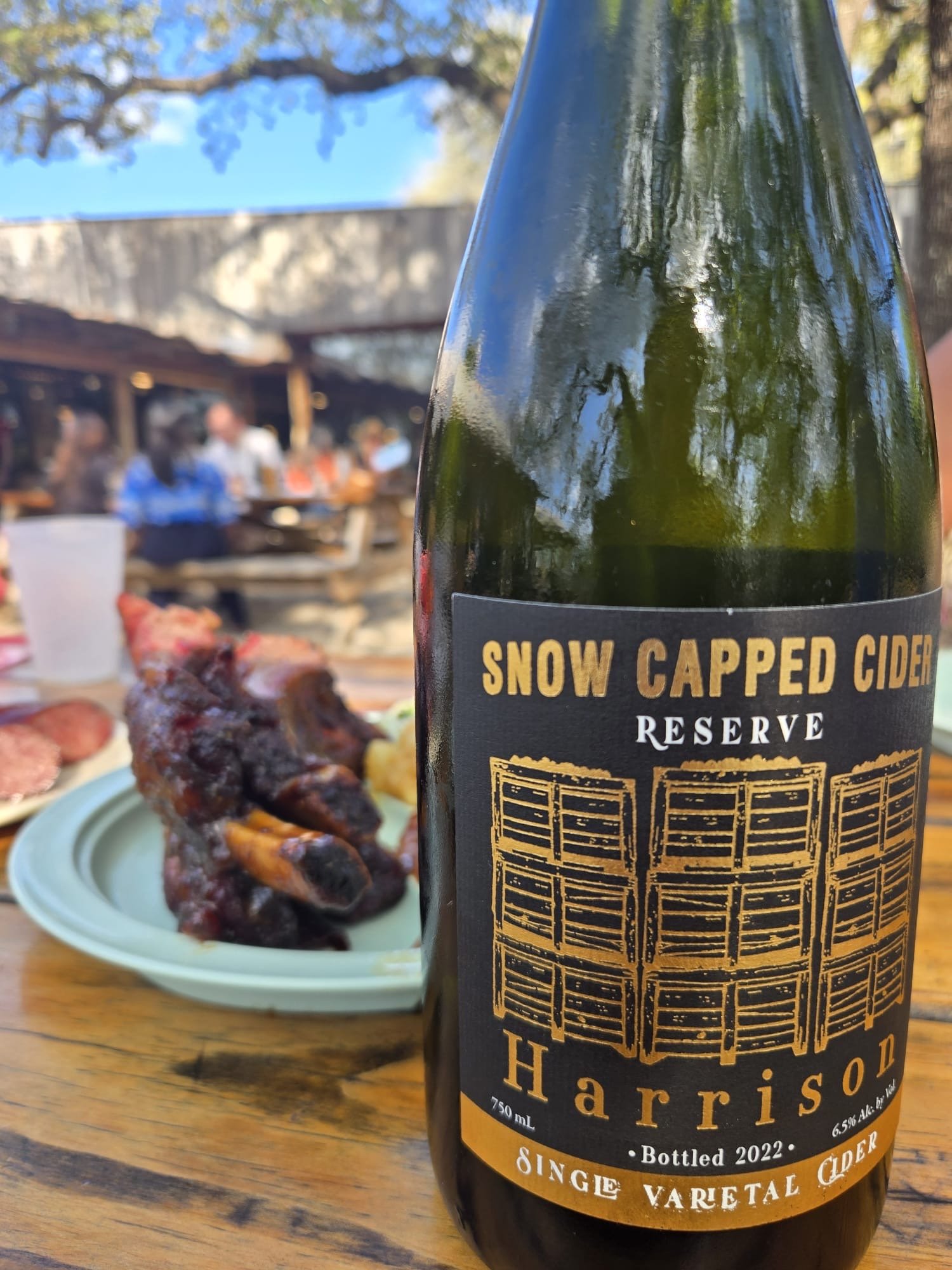 A bottle of Snow Capped Cider “Sparkling Rosé Cider” made with apples and elderberries sits on a table beside a stemmed glass filled with a deep pink beverage.