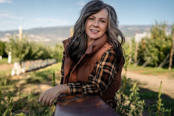 Multi-award-winning producer Kari Williams eans on a wooden fence in an apple orchard, wearing a brown vest and plaid shirt, with rows of trees and hills in the background at Snow Capped.
