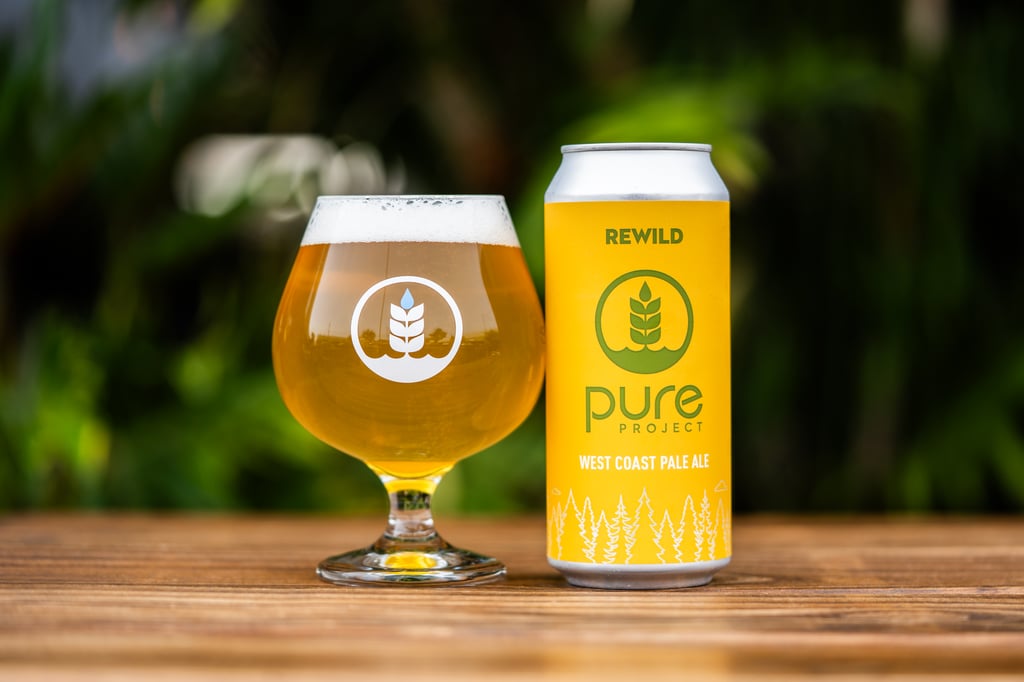 A tulip glass of golden pale ale beside a bright yellow Pure Project Rewild can on a wooden table
