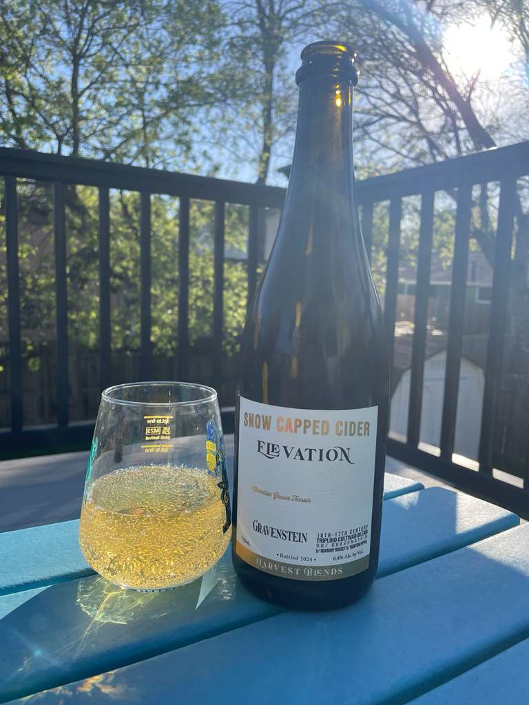 A bottle of Snow Capped Cider “Elevation” Gravestein cider stands on an outdoor table beside a glass filled with a golden, lightly sparkling drink.