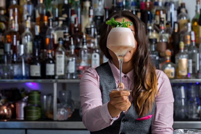 A bartender holds up a unique, creamy cocktail garnished with fresh mint in front of a well-stocked bar.