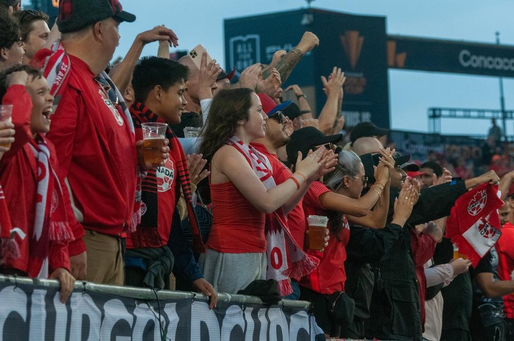 Fans in red cheer and clap in a stadium, building excitement just before kickoff.