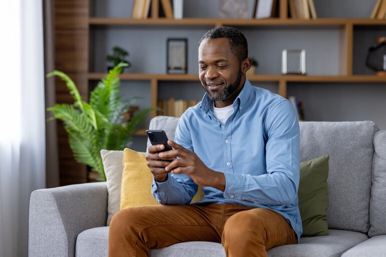 A man sitting on a living room sofa using a smartphone, representing the evolution of Melbet download across mobile and home devices.