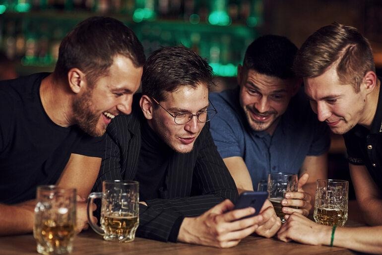 Friends having fun with the mobile phone in a beer garden, beers on the table, capturing sporting and group chat moments.
