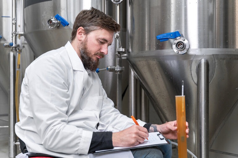 STEM student recording brewery production measurements beside stainless steel brewing tanks.