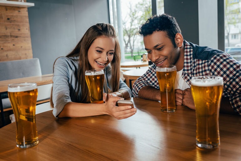 Couple on a date at a bar using a smartphone while drinking beer, highlighting how dating apps and AI could reshape real-world connections.