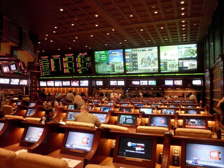 Sportsbook betting hall with electronic odds boards, betting screens, and gamblers placing sports wagers.