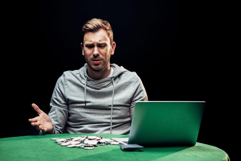 Worried man sitting in a casino with chips on the table, looking at his laptop screen showing the casino status as it loses its licence.