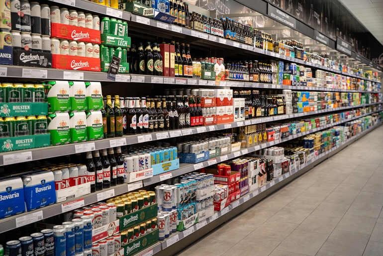 A supermarket beer aisle stocked with six-packs and cases of beer, illustrating why the same favorite six-pack can cost more in another state.