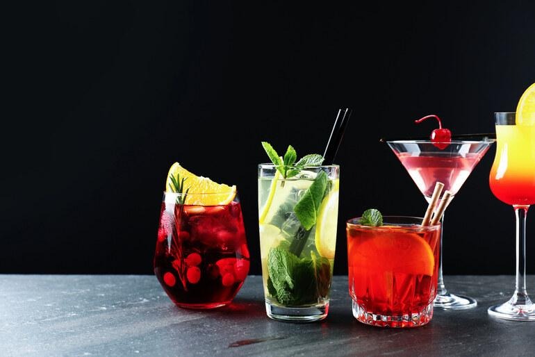 A row of various colorful cocktails including a mojito, martini, and fruit punch served on a dark bar surface.