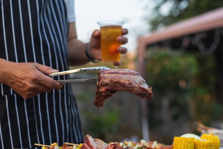 Man grilling steak at a backyard BBQ while preparing classic food and pairing drinks for an outdoor meal.