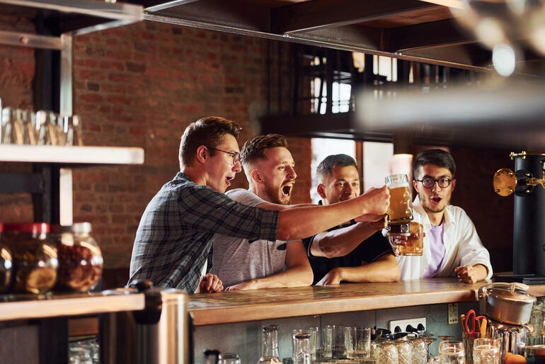 Four men sitting at a bar, celebrating with beer in their hands.