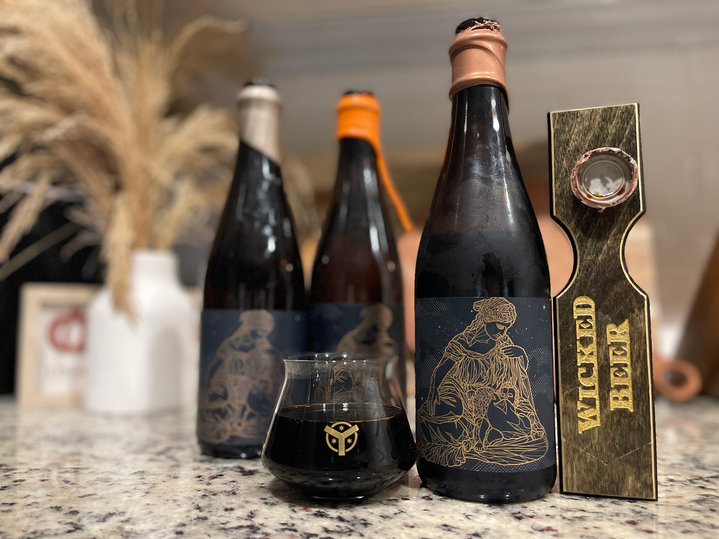 Review Image of Double Barrel Leto Beer. Small taster glass, two other variations of Leto stout in the background. Accompanied by Three Branch Studio custom wooden opener.