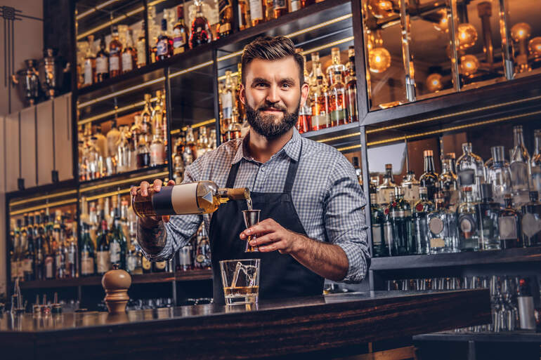 A stylish bartender in a shirt and apron is making a cocktail at a bar