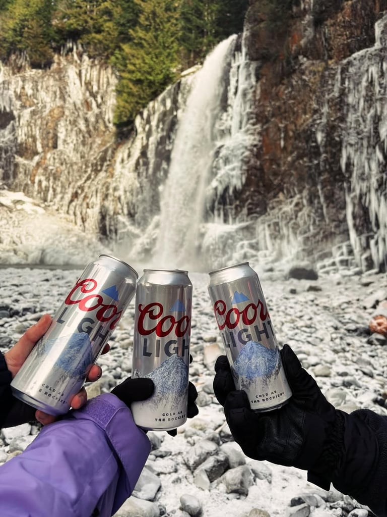 Three people in winter gear holding up tall cans of Coors Light for a toast, with a frozen waterfall and rocky, snow-covered terrain in the background.
