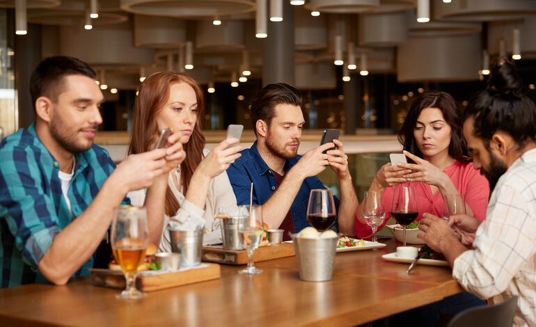 Friends at a restaurant focus on their phones instead of each other, reflecting Ireland’s shift from local social spaces to online interaction.